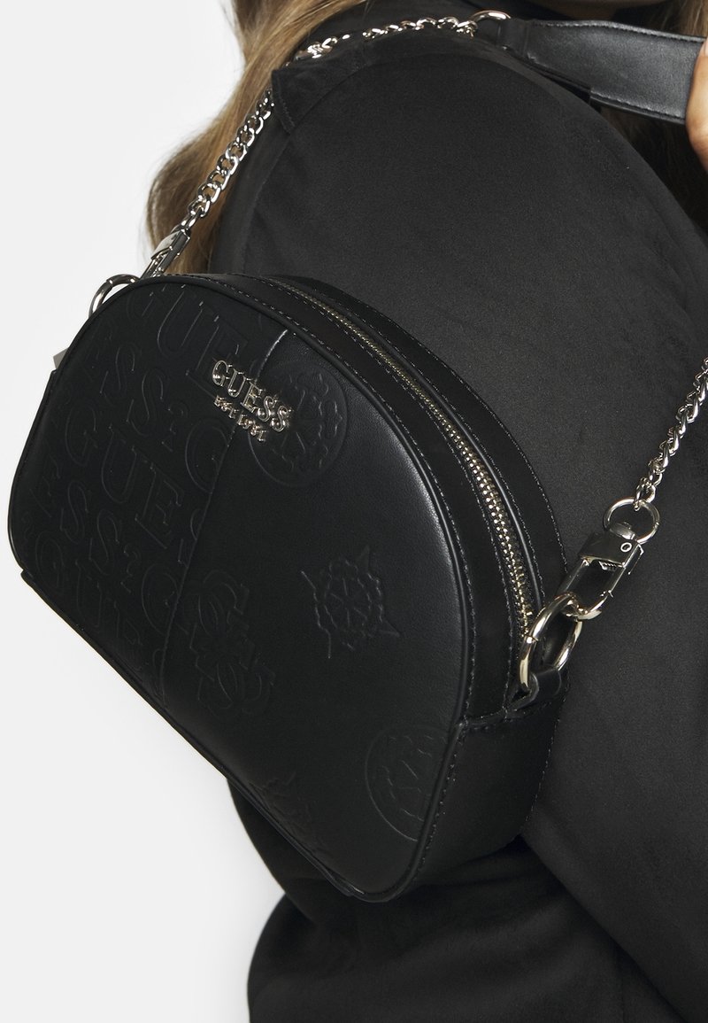 Sac à main noir Guess en forme de demi-lune avec motif de logo embossé et bandoulière chaîne argentée porté sur l'épaule avec des vêtements noirs.