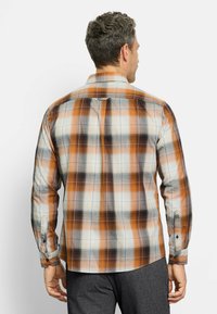 Mann mit kurzen lockigen Haaren, der ein langärmliges hemd in orange, weiß und grau im Karomuster trägt, von hinten vor einem weißen Hintergrund zu sehen.