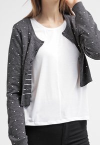 Šedý cardigan s bílými puntíky, pletená textura, zapínání na knoflíky a pruhovaný design na vnitřní légě přes bílou košili.