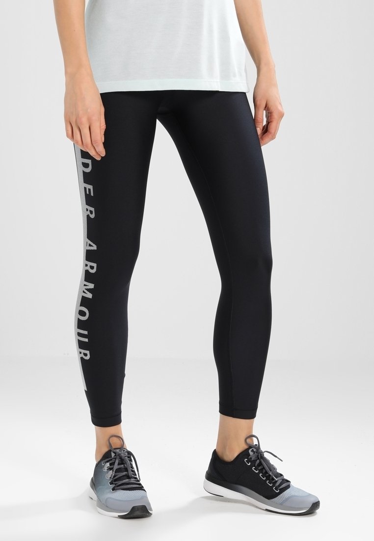 Leggings neri con il testo riflettente "Under Armour" sul lato, realizzati in un materiale liscio ed elastico. Abbinati a scarpe da ginnastica grigie.