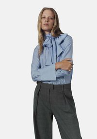 Blusa azul claro con un gran lazo en el cuello, combinada con pantalones oscuros a rayas grises con cintura alta y pliegues.