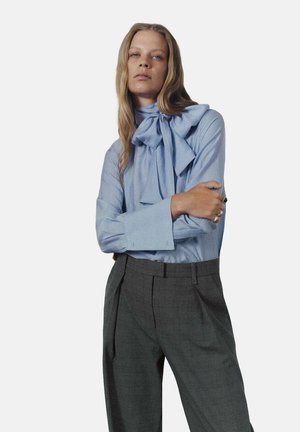 Blusa azul claro con un gran lazo en el cuello, combinada con pantalones oscuros a rayas grises con cintura alta y pliegues.
