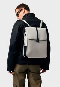 Grey backpack with flap closure, contrasting black strap, and textured fabric. Features minimalistic design and front mesh pocket.