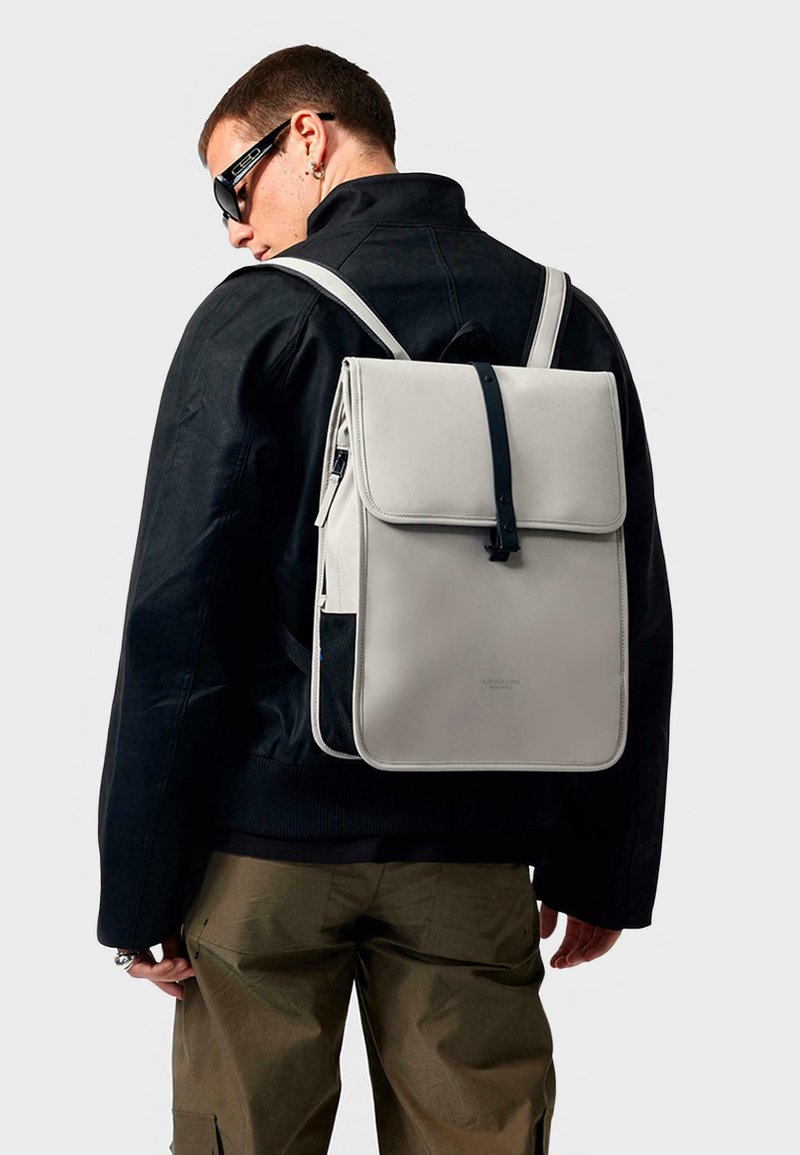 Zaino grigio con chiusura a ribalta, cinturino nero a contrasto e tessuto testurizzato. Presenta un design minimalista e una tasca frontale in rete.