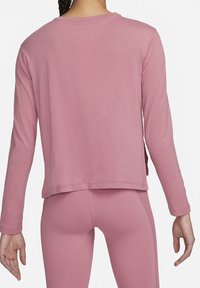 Frau in einem staubigen rosa Langarmoberteil und passenden engen Leggings, von hinten gezeigt, mit sichtbarem geflochtenem Haar.