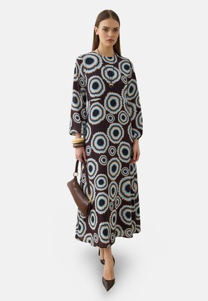 Donna che indossa un vestito nero a maniche lunghe con motivi circolari blu e bianchi, tiene una borsa marrone, indossa braccialetti e scarpe con punta.
