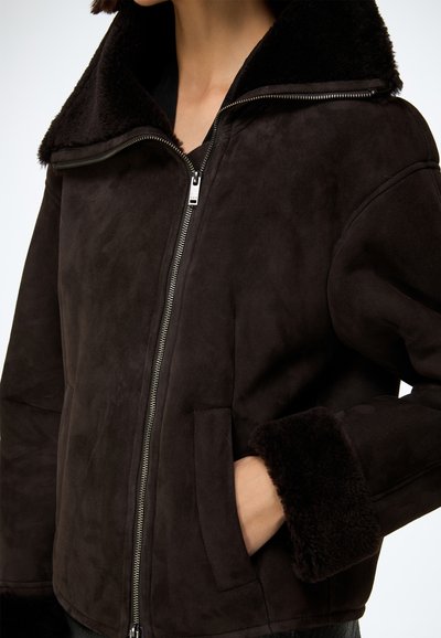 Brązowa zamszowa kurtka z dużym, miękkim czarnym kołnierzem i mankietami z owczej skóry. Posiada przedni zamek błyskawiczny oraz dwie boczne kieszenie. Przyjemna w dotyku.