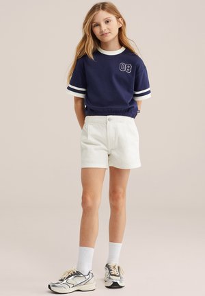 Fată tânără cu păr blond lung, purtând un tricou scurt bleumarin cu inscripția „08”, pantaloni scurți albi, șosete albe și pantofi sport argintii, stând în picioare.