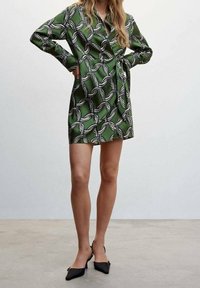 Grünes gemustertes Wickelkleid mit schwarzem und weißem abstraktem Design, langen Ärmeln und einem Bindegürtel, kombiniert mit schwarzen spitz zulaufenden Absätzen.
