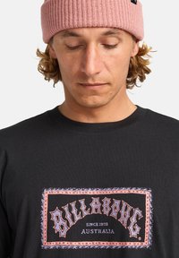 Sort t-shirt med et stort grafisk motiv, der viser ordet "BILLABONG" i stiliseret orange og lilla tekst, båret med en pink strikket beanie.