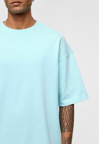 T-shirt surdimensionné bleu clair en coton doux, avec un col rond et des manches courtes. Tatouage visible sur le bras.