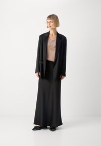 �Černý oversized blazer přes béžový top se zlatou výšivkou, doplněný dlouhou černou sukní a černými botami se dekorativními akcenty.