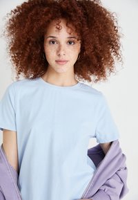 Lyseblå bomulds t-shirt med korte ærmer og rund hals, lagd under en lavendeljakke med lynlås og ribbede manchetter.