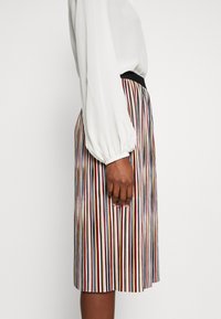 Une jupe plissée jusqu'aux genoux avec des rayures verticales multicolores, dotée d'une taille élastique noire, associée à un haut blanc à manches longues.