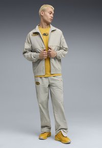 Lys grå sporty jakke med elastiske manchetter, parret med en sennepsgul t-shirt og matchende lys grå bukser, der har logo detaljer og gule sko.