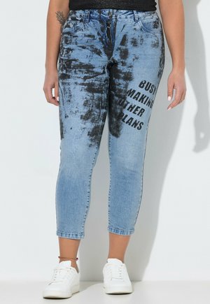 Underkrop iført blå og sort tie-dye jeans med teksten "Busy making other plans" på højre lår, kombineret med hvide sneakers.