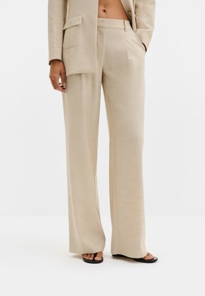 Beige breite Anzughose mit Fronttaschen, getragen mit passendem Blazer und schwarzen Sandalen mit quadratischer Spitze an einem Modell.
