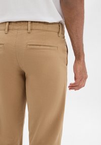 I chinos color khaki presentano una texture liscia, un taglio dritto e due tasche posteriori. Una mano è visibile, appoggiata in modo casual sul coscia.