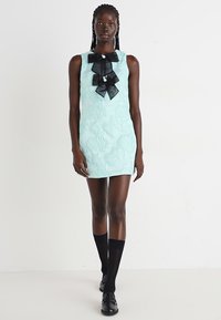 Minzgrünes ärmelloses Kleid mit strukturiertem Blumenmuster, schwarzer Schleifenakzent am Ausschnitt, kombiniert mit kniehohen schwarzen Socken und Slippern.