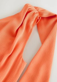 Haut sans manches en tissu texturé corail-orange avec un détail noué au niveau du col sur fond blanc.