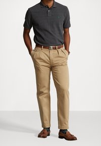 Mörkgrå pikétröja med kort ärm, grön logotyp på bröstet, ihopparad med beige chinos, brunt bälte och bruna loafers.