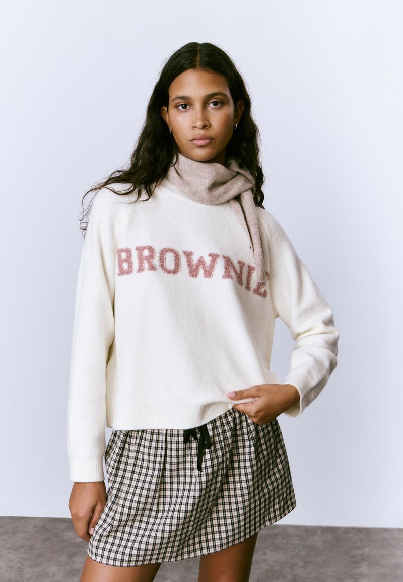 Suéter crema con texto "BROWNIE" en rosa, combinado con una bufanda beige y una falda a cuadros en blanco y negro. Material suave y corte holgado.