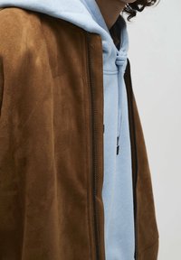 Bruine suède jas met een slank design en een zwarte rits, gelaagd over een lichtblauwe hoodie met een trekkoord. Zachte textuur is duidelijk zichtbaar.