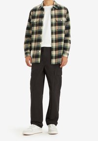 Man i grön och beige rutad skjorta med knäppning över vit t-shirt, svarta cargo-byxor och vita sneakers, stående mot vit bakgrund.