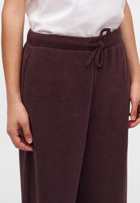 Gros plan d'une personne portant un pantalon en velours côtelé violet foncé avec une taille à cordon nouée et une chemise blanche rentrée.