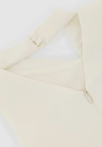 Fehér ruhaszövet sima textúrával, V-nyakú kialakítással és oldalsó cipzárral. Két kerek gomb díszíti a vállnál.