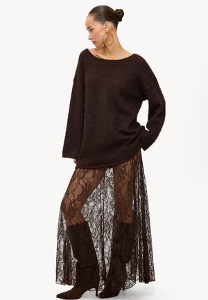 Mujer vistiendo un suéter de gran tamaño de color marrón oscuro y peludo, una falda larga de encaje negro transparente, botas marrón hasta la rodilla y grandes pendientes de aro dorados, de pie de lado.