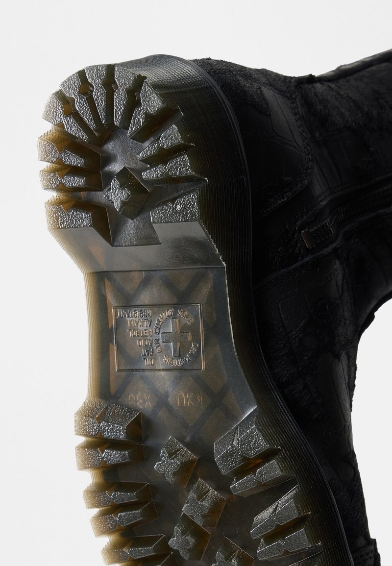 Suola di uno stivale nero con battistrada in gomma profonda, cerniera laterale e simbolo a croce con testo sul fondo traslucido.