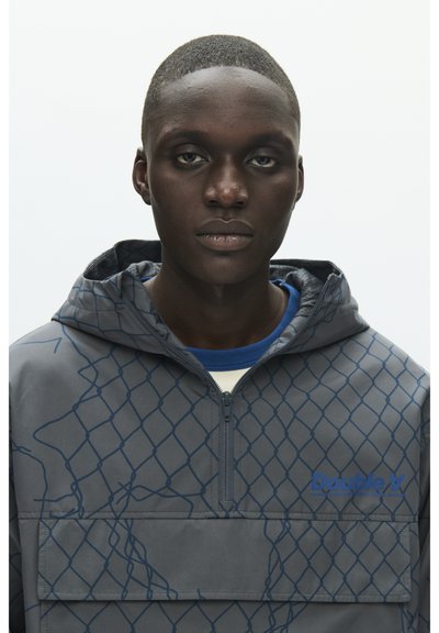 Jeune homme noir portant une veste à capuche grise zippée avec un motif géométrique bleu en grille et une chemise bleue en dessous.
