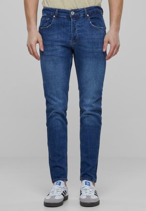 Modre jeans hlače z ožjim krojem, petimi žepi in gumbom za zapiranje. Poudarjene so s svetlim bledenjem in šivnimi detajli. V kombinaciji z belimi športnimi copati.
