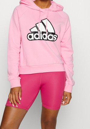Osoba ubrana w jasno różową bluzę z kapturem Adidas i jaskrawo różowe dopasowane szorty, stojąca na tle jednolitego jasnego tła.