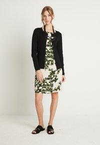 Cardigan nero su un vestito crema con motivi di foglie verdi, abbinato a sandali neri. Collana di perline con accenti neri.
