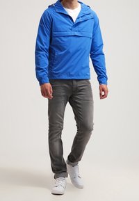 Blå lätt anorak med halvdragkedja fram och känguruficka. Matchat med grå jeans och vita sneakers. Slät textur, avslappnad passform.