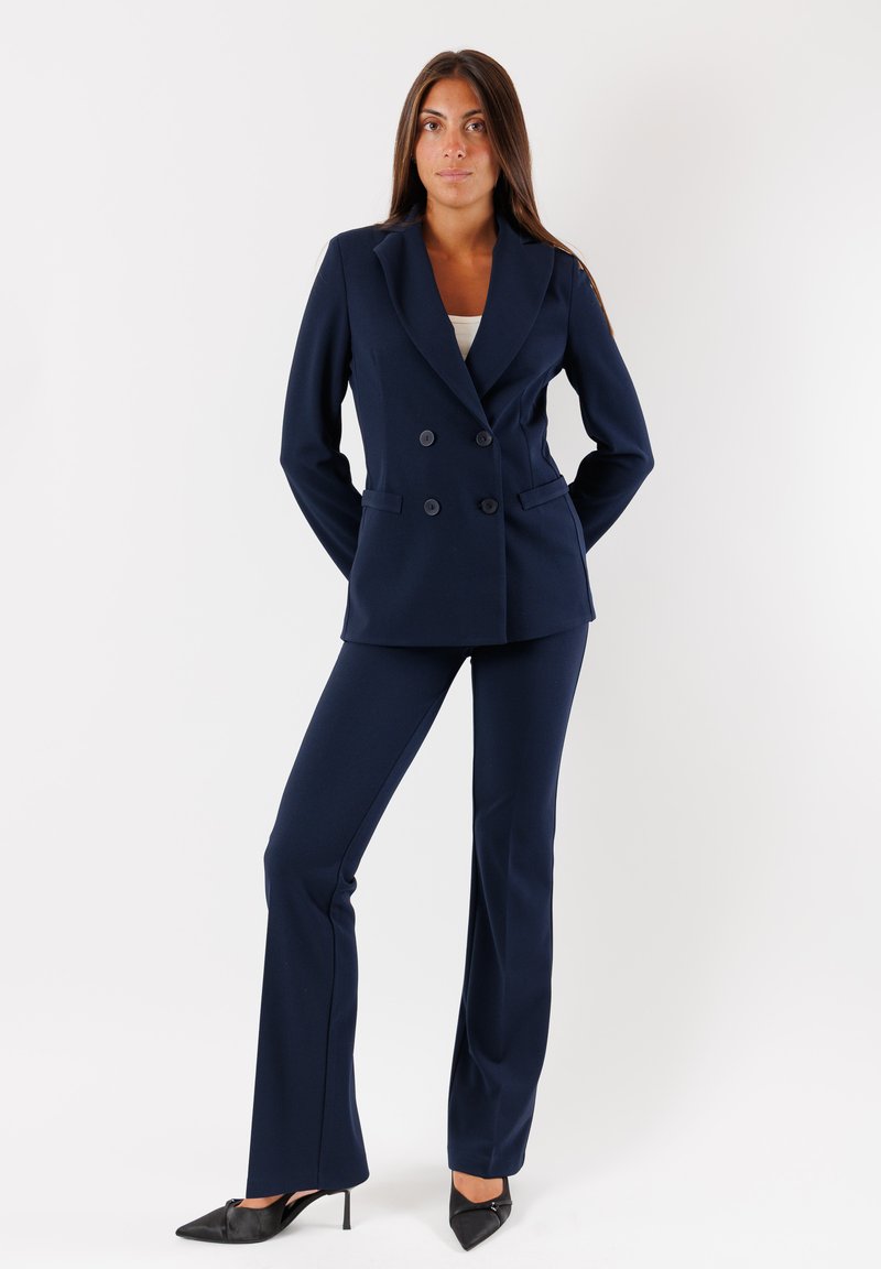 Blazer doppiopetto blu marina con rever, abbinato a pantaloni a campana coordinati. Il materiale appare liscio e ben sartoriale. Tacchi neri a punta.
