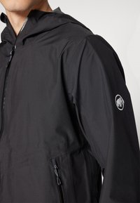 Veste légère noire avec capuche, dotée d'une fermeture éclair, de poches latérales et d'un logo d'éléphant réfléchissant sur la manche. Tissu texturé.