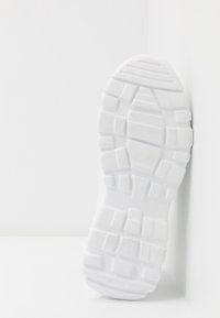 Semelle de chaussure blanche avec un motif de crampons profonds et carrés, inclinée contre un mur et un sol blancs dans un cadre minimaliste.