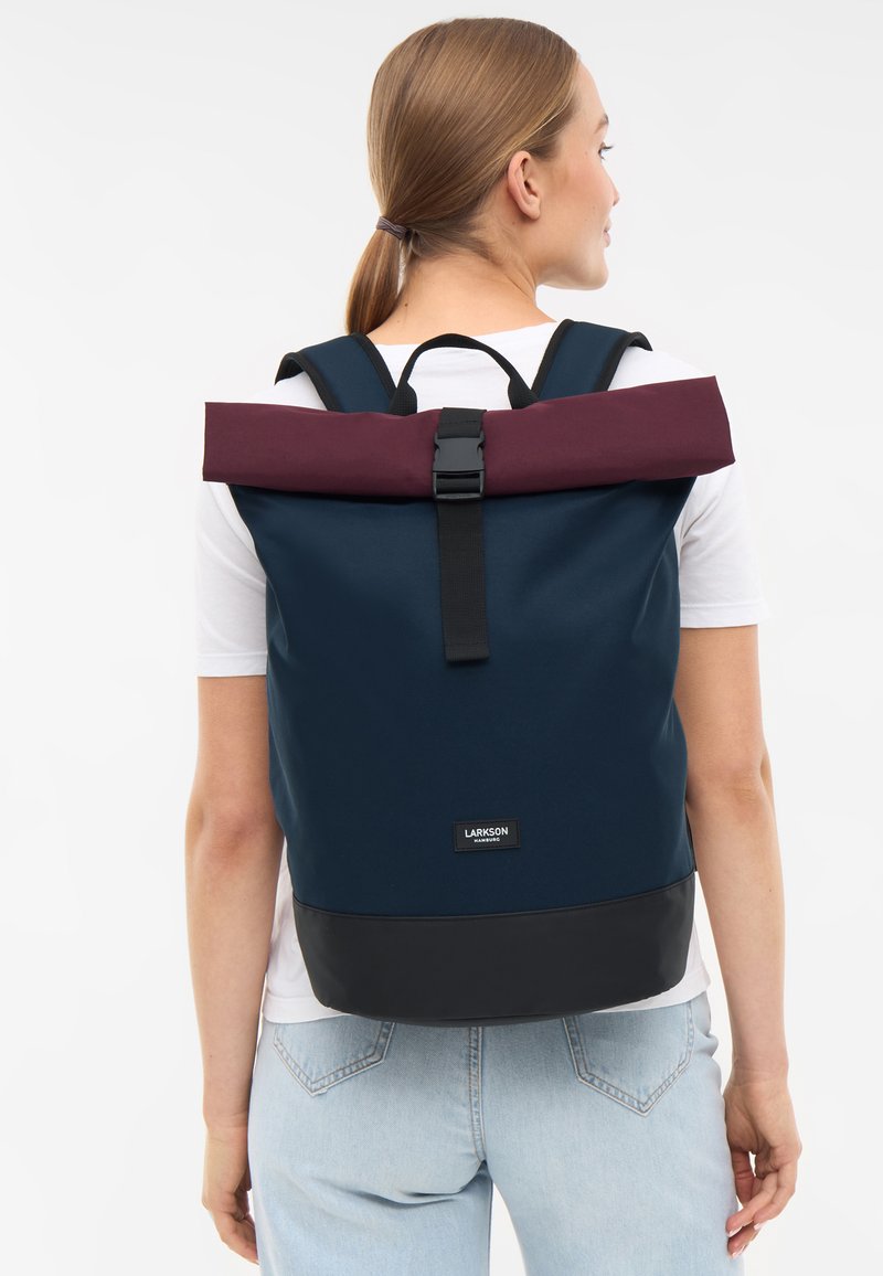 Tengerészkék roll-top hátizsák bordó kiemelésekkel, fekete alappal, állítható pántokkal és elöl csattal záródik. Simább textúra, modern dizájn.