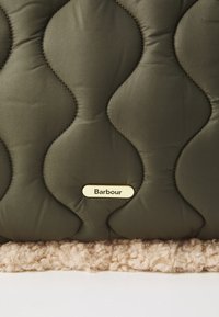 Kváltott olívazöld szövet hullámos varrásokkal, puha bézs plüss szegéllyel és egy arany fém címkével, amelyen "Barbour" felirat szerepel.