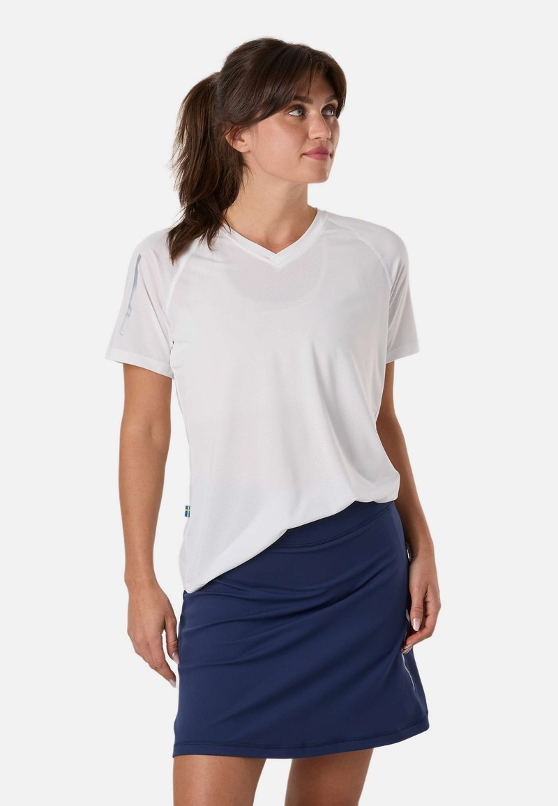 Femme aux cheveux bruns en queue de cheval portant une chemise blanche ample à manches courtes et une jupe bleu marine, regardant vers sa gauche sur un fond blanc.