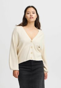 Ljust beige ribbat cardigan med V-ringning, långa ärmar och fyra knappar framtill. Bärs över en svart kjol, texturerad tyg.