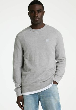 Mann mit dunkler Haut, der einen hellgrauen Strickpullover über einem weißen Hemd und hellblauen Jeans trägt, steht mit einer Hand in der Tasche.