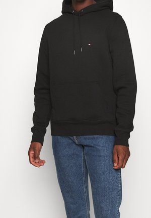 Person trägt einen schwarzen Hoodie mit Kordelzug und kleinem Logo auf der Brust, kombiniert mit blauen Jeans, steht vor einem schlichten Hintergrund.