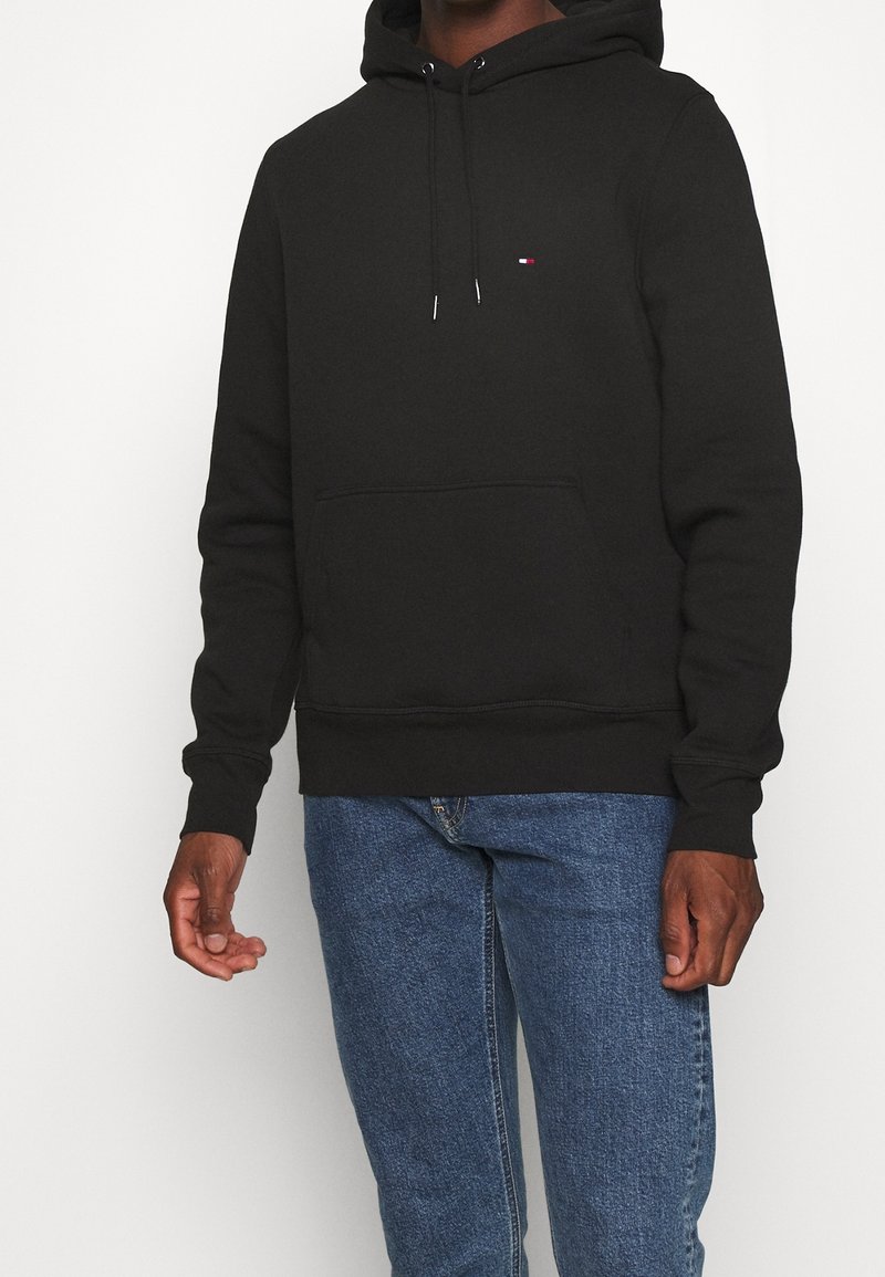 Personne portant un sweat à capuche noir avec des cordons et un petit logo sur la poitrine, associé à un jean bleu, debout devant un fond uni.