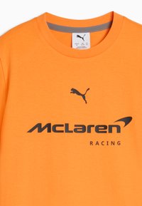 T-shirt à col rond orange vif avec logo Puma noir au-dessus du texte noir McLaren Racing au centre avant.