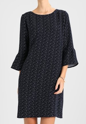Femme portant une robe bleu marine à petits pois blancs avec des manches cloche trois-quarts, debout devant un fond uni clair.