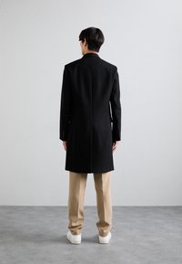 Abrigo de lana negro con corte recto, cuello y una abertura trasera, llevado sobre pantalones beige y zapatillas blancas, visto desde detrás.